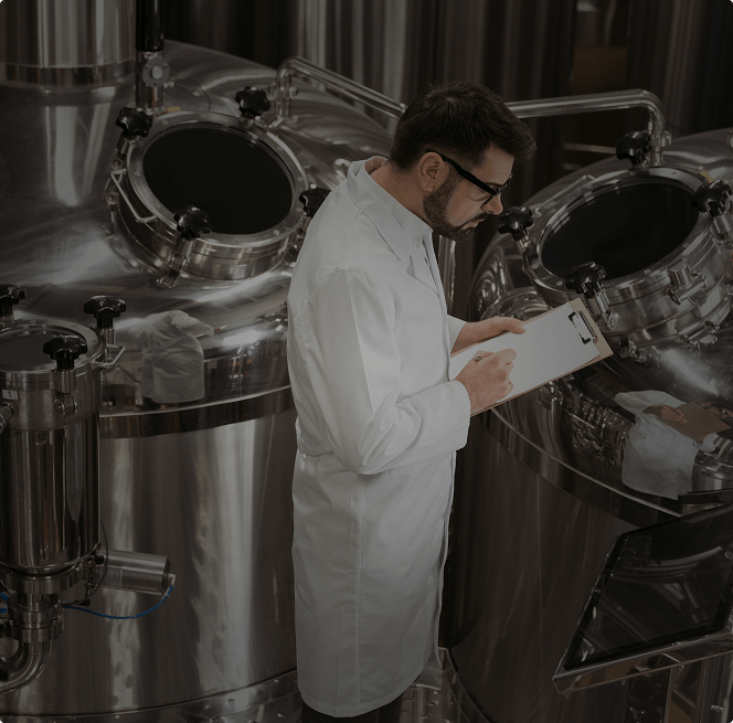 Lab-coated technologist inspecting stainless steel distillation vessels with sight glasses