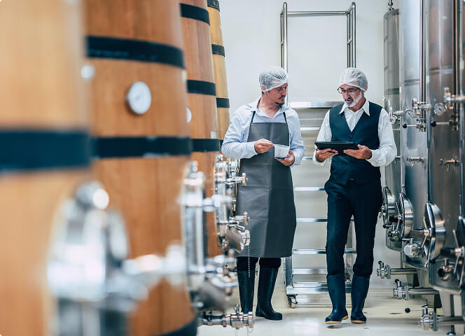 Two production specialists reviewing documentation beside barrels and fermentation tanks