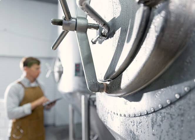 Closed hatch of a stainless steel tank with a worker holding a tablet in the background