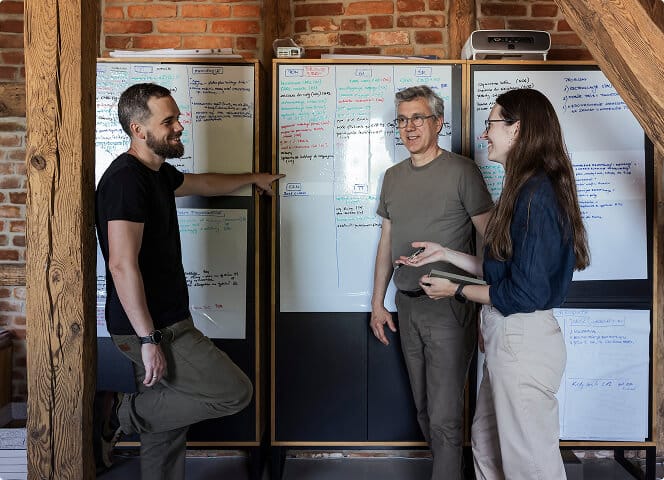 Three Alquora employees talking by whiteboards full of notes in a brick room with wooden beams