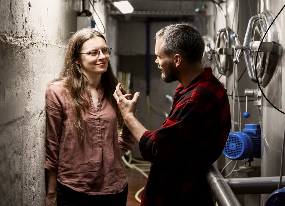 Two production specialists reviewing documentation beside barrels and fermentation tanks